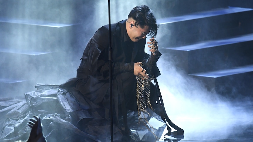 An emotional young man in a black gown crouches down and puts his hand over his face. 
