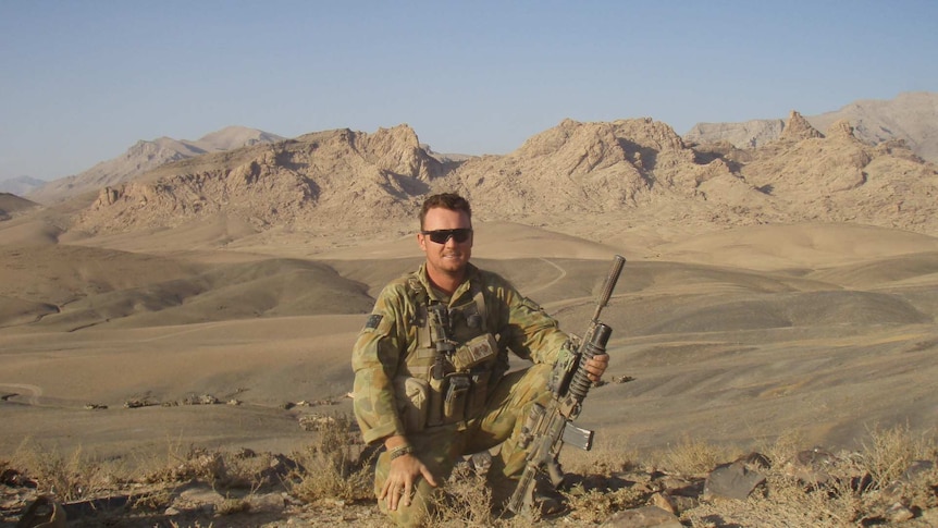 Mick Bainbridge, with gun in hand, sits in a desert while on a deployment with the Australian Defence Force
