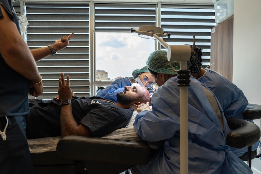 A man lays down on a hospital bed while chatting to four medical staff in scrubs who are inspecting his scalp