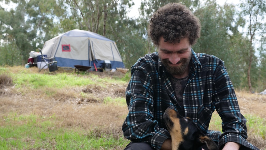 A man looks down at his dog in a jacket with a tent in the background on a hill.