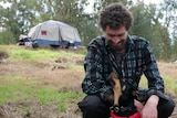 A man looks down at his dog in a jacket with a tent in the background on a hill.