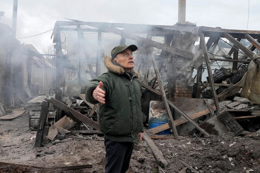 A man opens his arms as he stands near the charred wreckage of a house destroyed by artillery shelling.