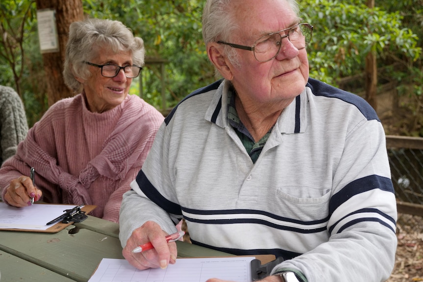 Man and woman sitting at table in a bush setting with score sheets on clipboards