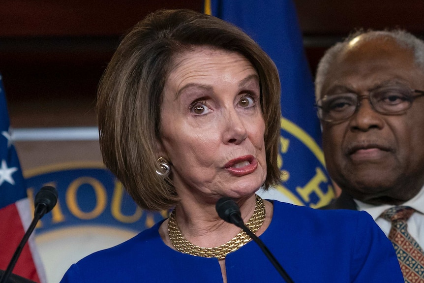 Nancy Pelosi wearing a blue top speaks at some microphones.