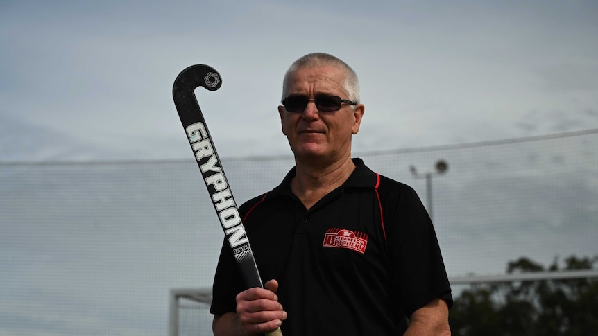 A man stands with a serious expression on his face holding a hockey stick over his shoulder on an empty field.