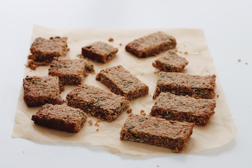 Rectangular pieces of a muesli bar slice sit randomly spaced on a piece of baking paper.