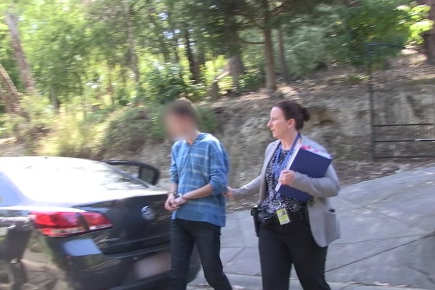 An arrested man with face obscured is led to a car by an officer.