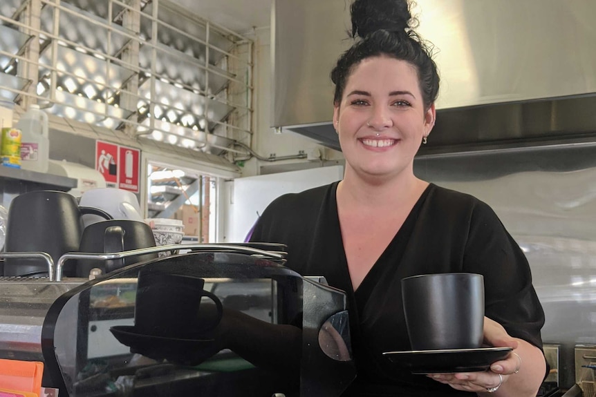 A woman dressed in black stands behind a coffee machine, smiling at the camera while holding out a black cup on a black saucer.