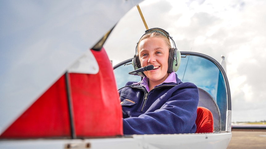 A girl sits in an open cockpit with a headset on grinning.