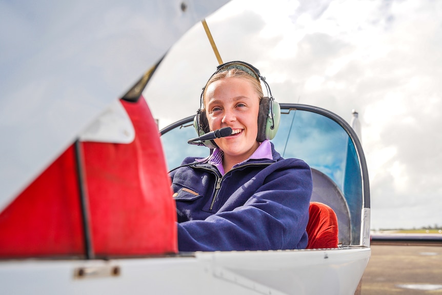 A girl sits in an open cockpit with a headset on grinning.
