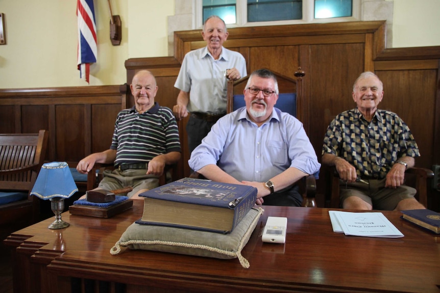 Four older men sitting on chairs in front of a desk inside mason hall.