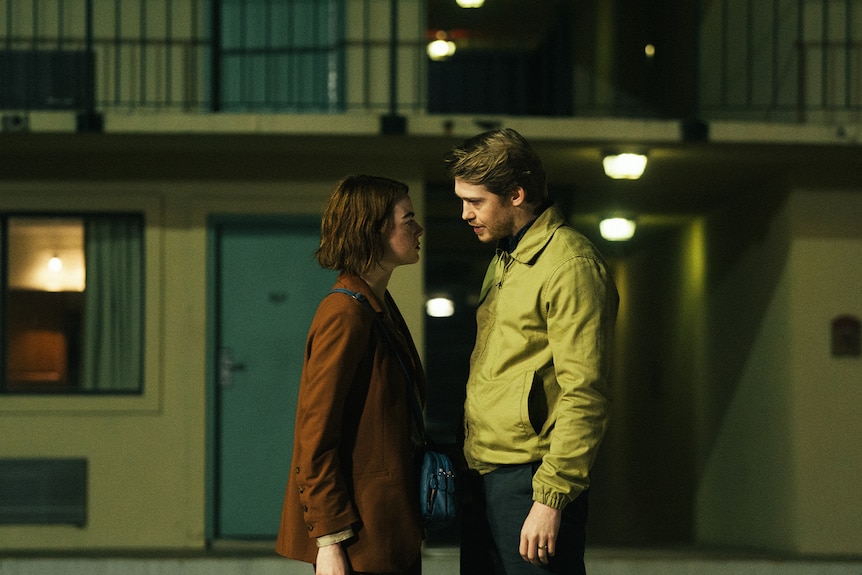 A man and a woman face one another, looking tense and standing close, in a dimly lit motel carpark at night.