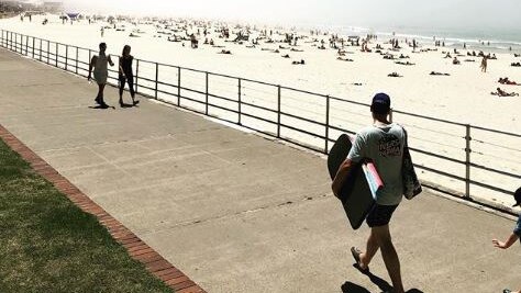 A man with a boogie board is seen walking along a footpath with Bondi beach in the background as fog rolls in.