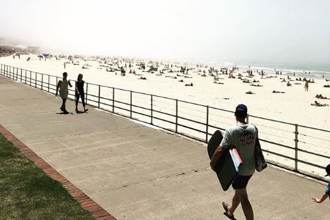 A man with a boogie board is seen walking along a footpath with Bondi beach in the background as fog rolls in.