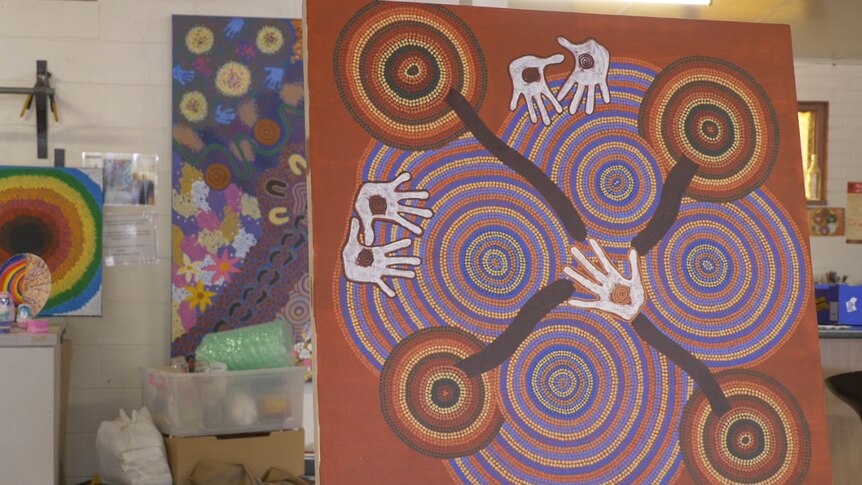 Photo of an earthy coloured dot painting on an easel, which features several hand prints.