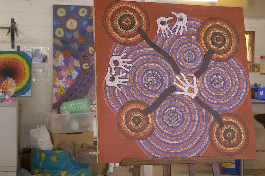 Photo of an earthy coloured dot painting on an easel, which features several hand prints.