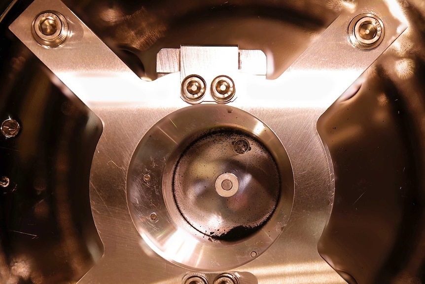 A small, dark sample of soil sits at the bottom of a gleaming metal container that is bolted into place