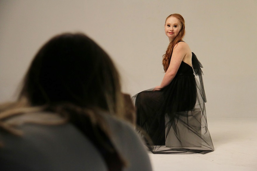 Maddie at a photoshoot posing on a chair in a black dress with photographer in foreground.