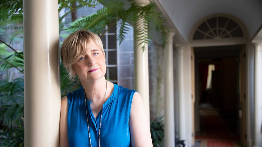A portrait of author Sue Williams standing in a courtyard, leaning against a column