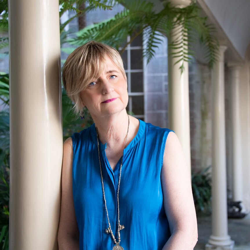 A portrait of author Sue Williams standing in a courtyard, leaning against a column