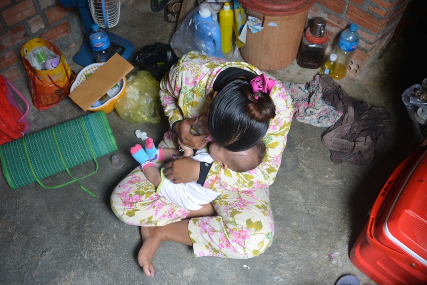 A woman in pink and green floral pyjamas pictured from above bottle feeding a baby in her lap.