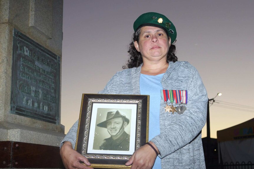 A woman poses for a photo wearing a beret and holding a picture of her grandfather in a soldier's uniform.