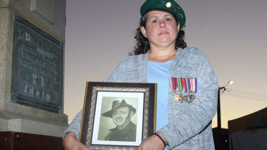 A woman poses for a photo wearing a beret and holding a picture of her grandfather in a soldier's uniform.