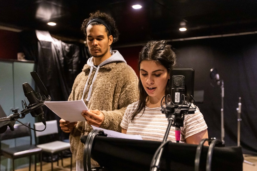 A tall young man with fluffy jacket stands next to young woman in sound recording studio, both in front of mics.