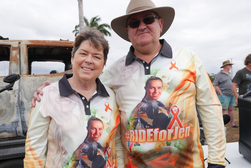 A man and a woman in matching shirts which read '#rideforJen' stand in front of a burned out car