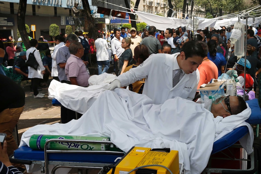 A doctor bends over a man lying in a hospital bed in the middle of a bustling street.