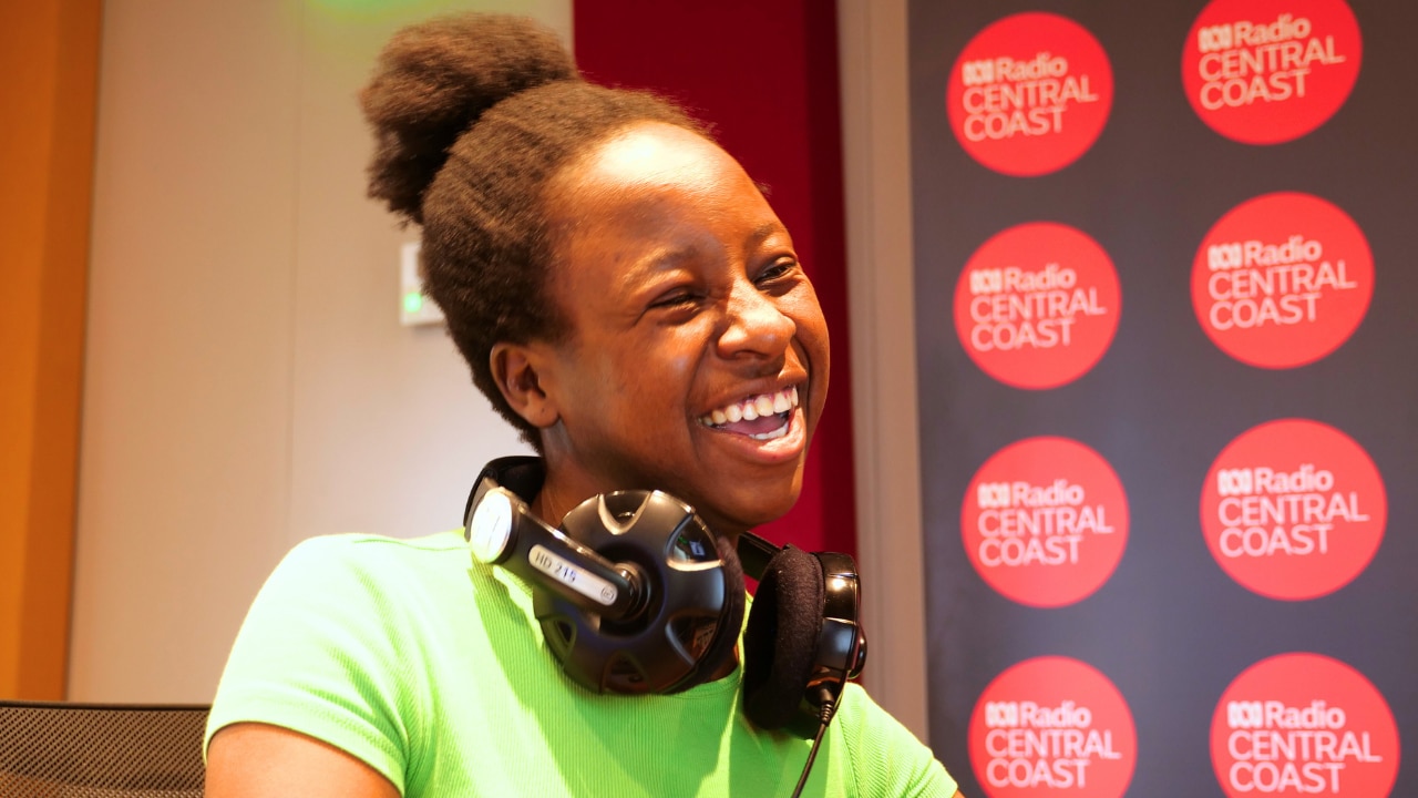 A young woman smiles. She wears headphones around her neck.