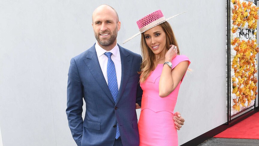 Chris Judd in a navy blue suits stands with his wife Rebecca in a pink dress and hat.