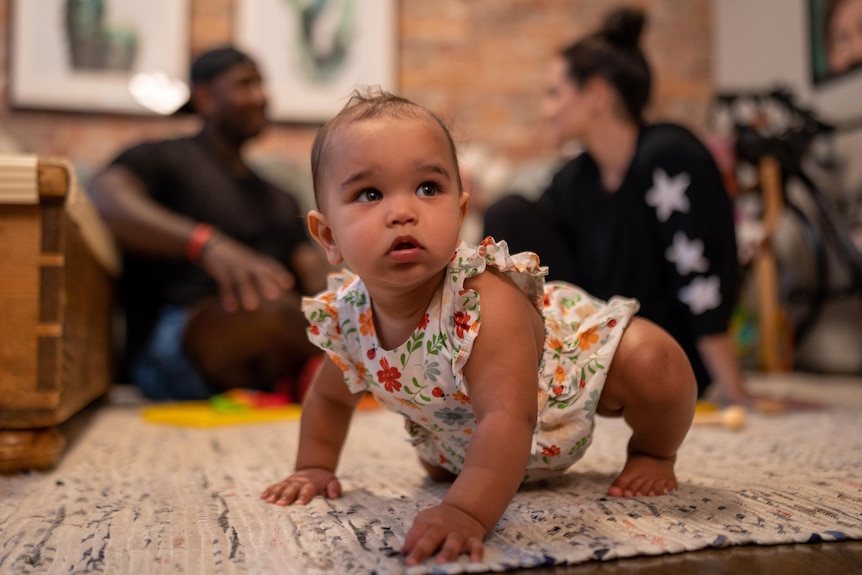 A baby in a floral jumpsuit crawls on a white rug while her parents chat nearby