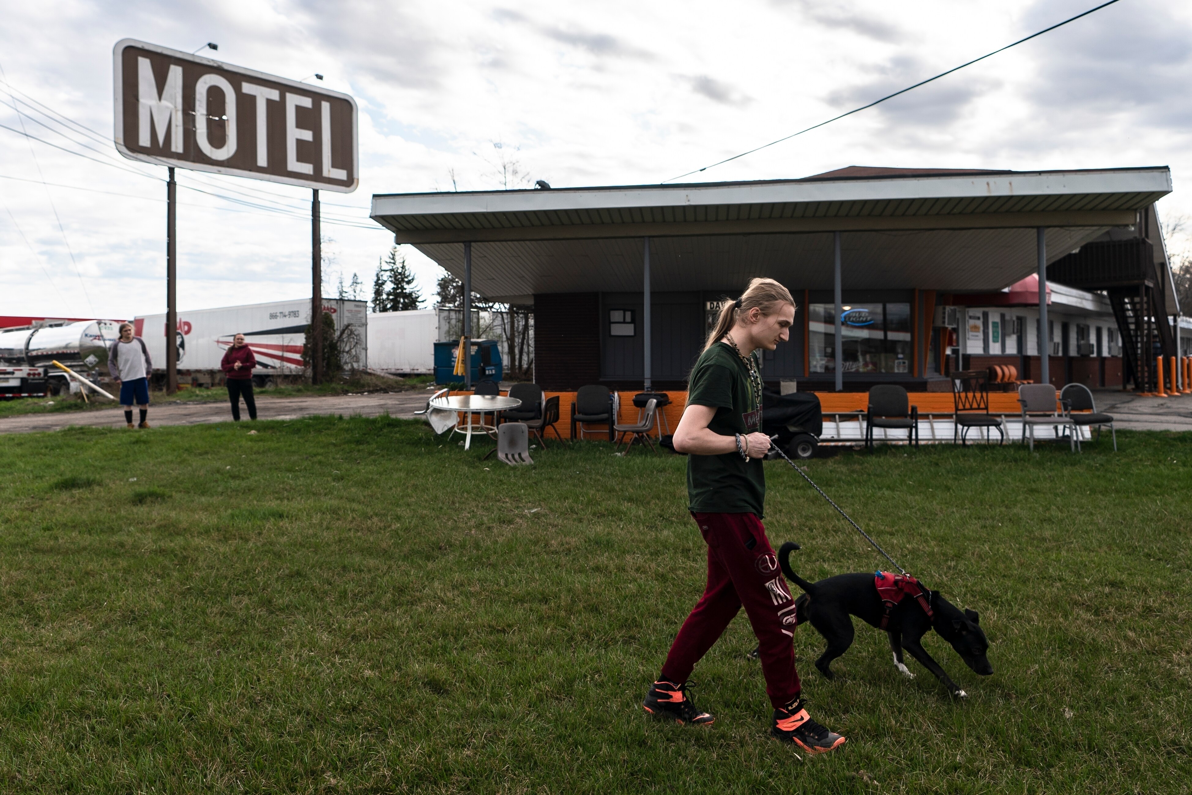 A young white man with long blonde hair in a ponytail walks a black dog on a patch of grass next to a motel.