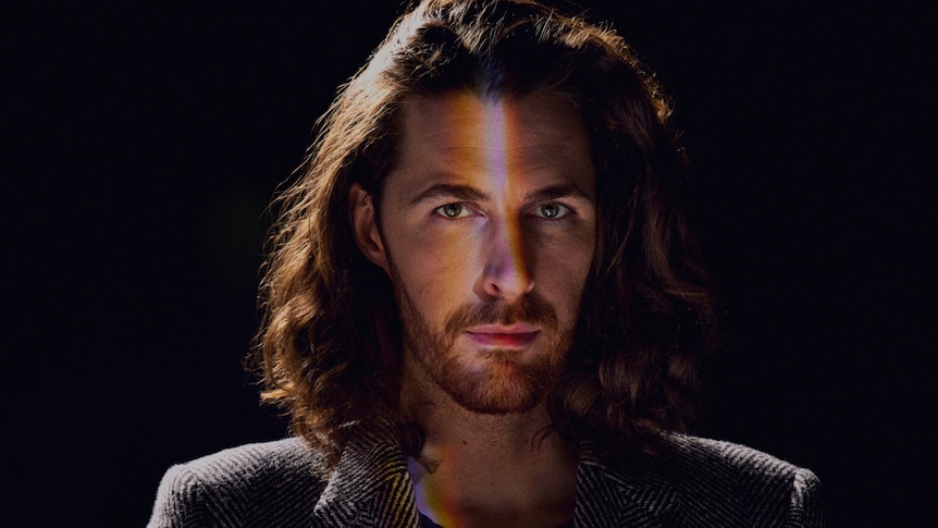 Hozier looks into the camera, coloured light reflects on his place. Blank dark background. 