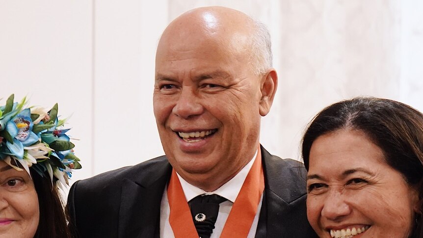 A pacific man with no hair smiling wearing a suit and with a medallion around his neck
