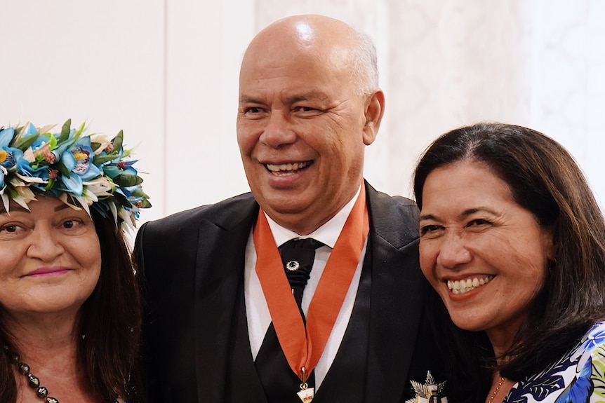 A pacific man with no hair smiling wearing a suit and with a medallion around his neck