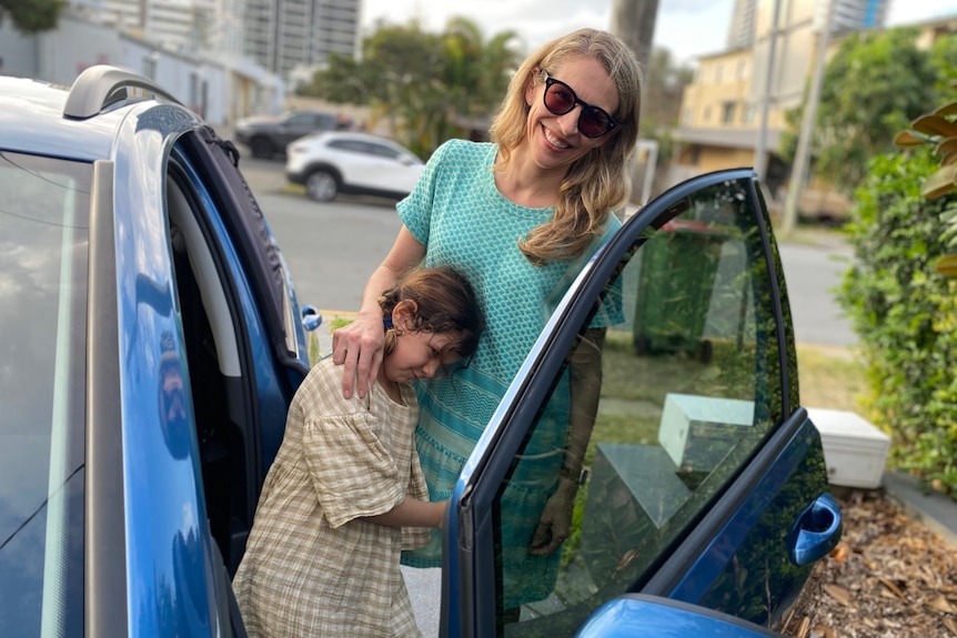 A woman and a child stand at the open door on the front passenger side of a blue car.