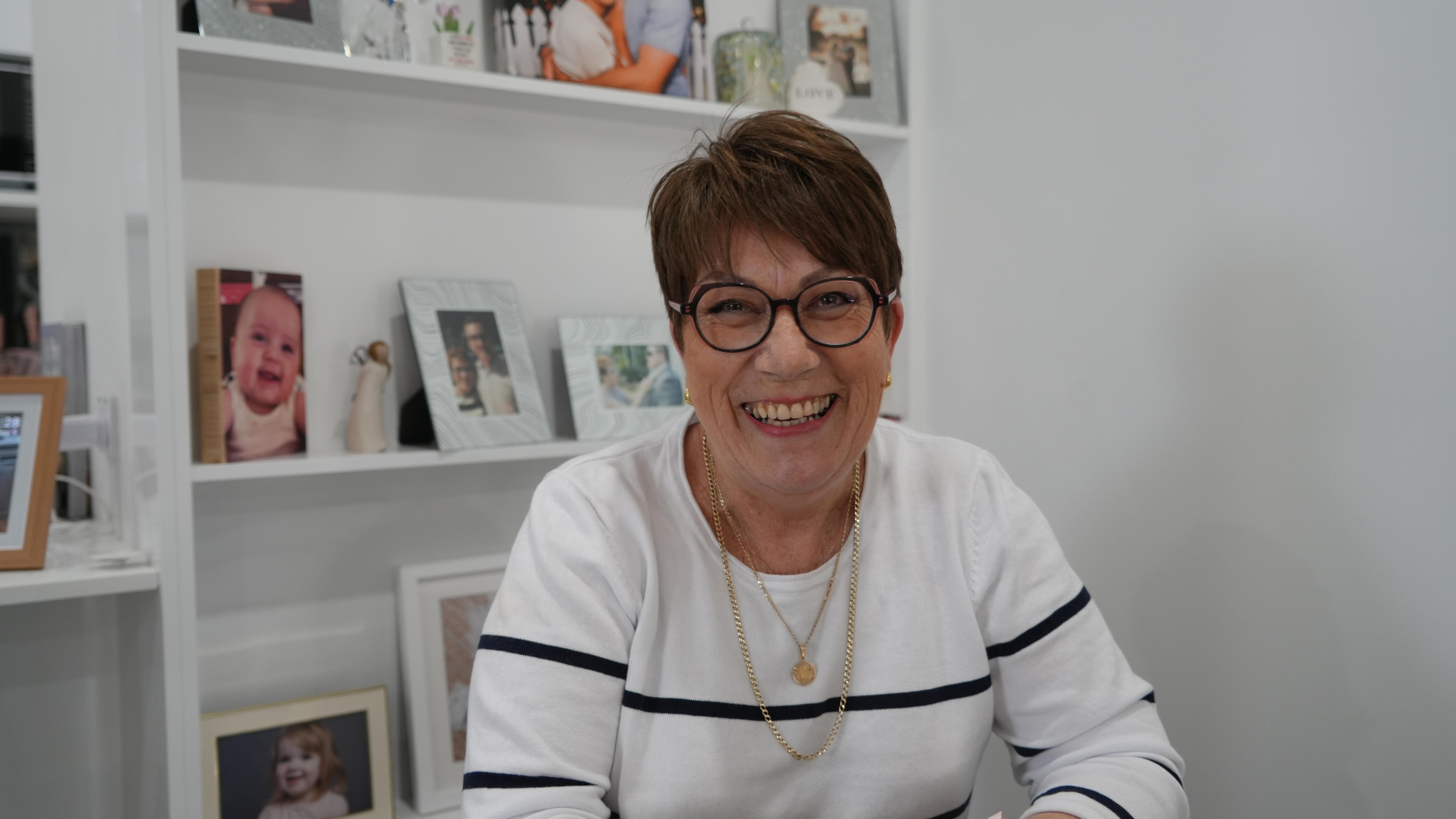 Smiling woman in white with stripe in front of photo frames on a shelf.
