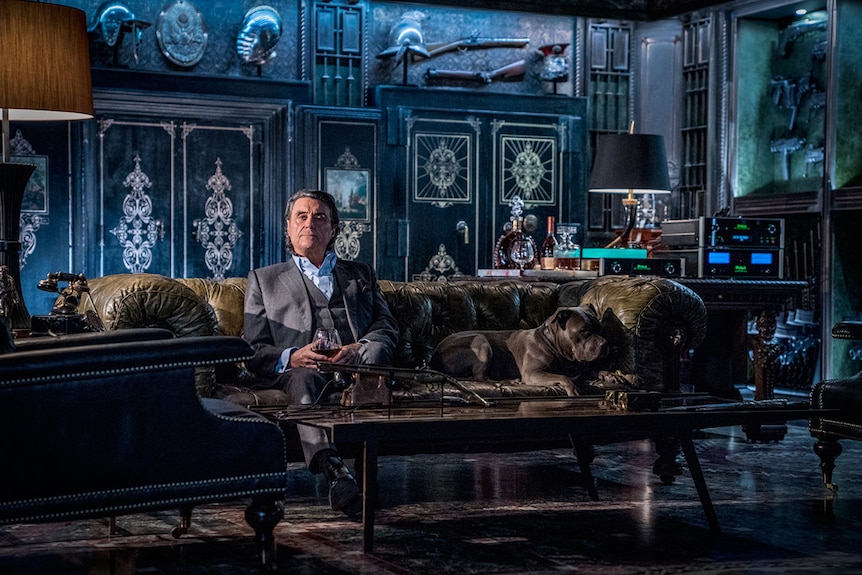 Colour still of Ian McShane sitting at on a sofa with a dog, in living room decorated with weapons in John Wick: Chapter 3.