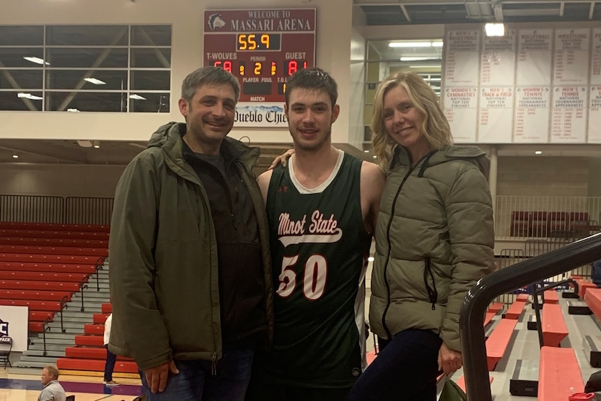 Young man holds smiling man, woman in green jackets inside a basketball. Young man wears basketball jersey with 50, Minot State.