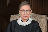 Ruth Bader Ginsburg in robes and a white collar sitting on an arm chair