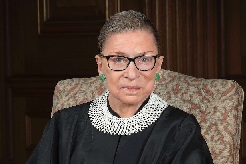 Ruth Bader Ginsburg in robes and a white collar sitting on an arm chair