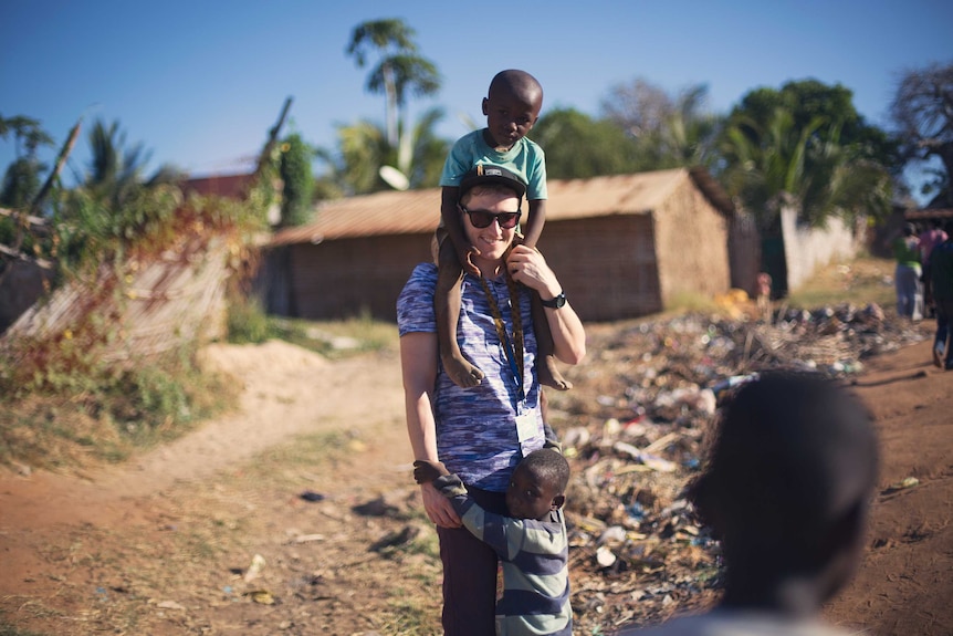 Tall Australian guy carrying a young African child on his shoulders, while another child grabs him round the legs.