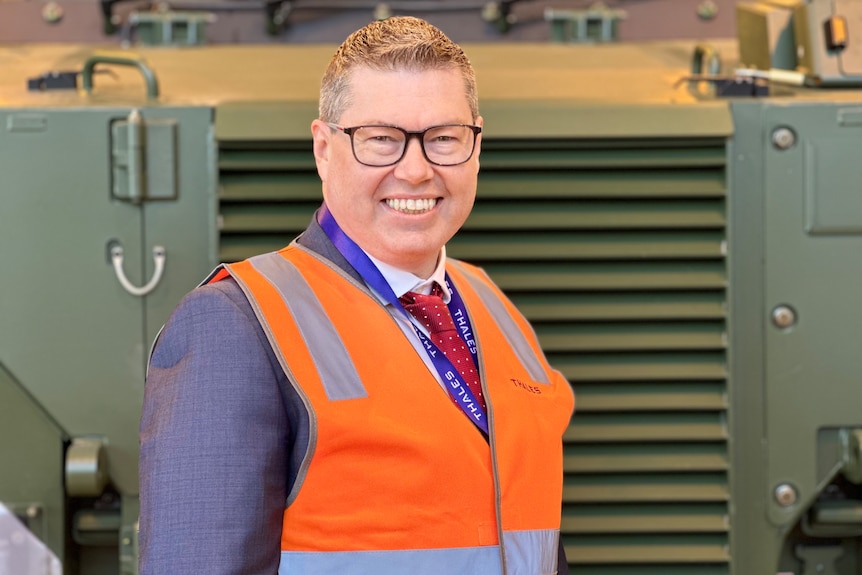 A man in glasses and a blue suit smiles at a camera while standing front of a Bushmaster