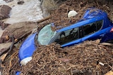 blue car covered in debris on the rocky shore of a beach
