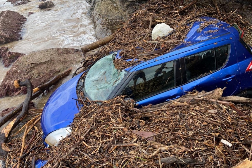 blue car covered in debris on the rocky shore of a beach