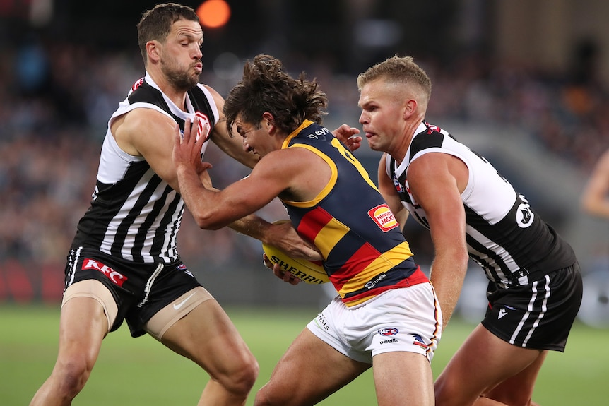 An Adelaide Crows AFL player holds the ball as he is tackled by two Port Adelaide opponents.