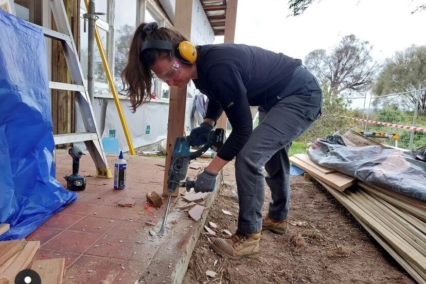 A woman in work wear and ear protection drilling some wood.