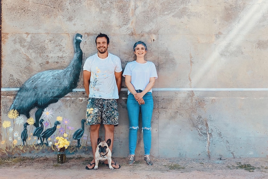 A man and woman with dog stand in front of a grey concrete wall painted with an emu and chicks